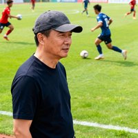 全国业余足球联盟秘书长赵刚，戴着棒球帽，站在绿茵场边，身后是正在比赛的业余球队球员